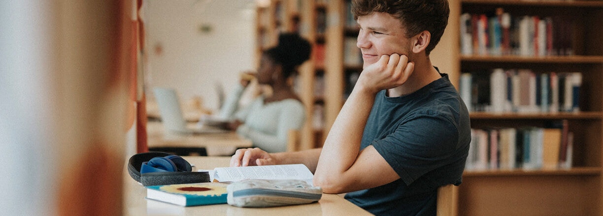 A student studying at a desk with books and a laptop in a library.
