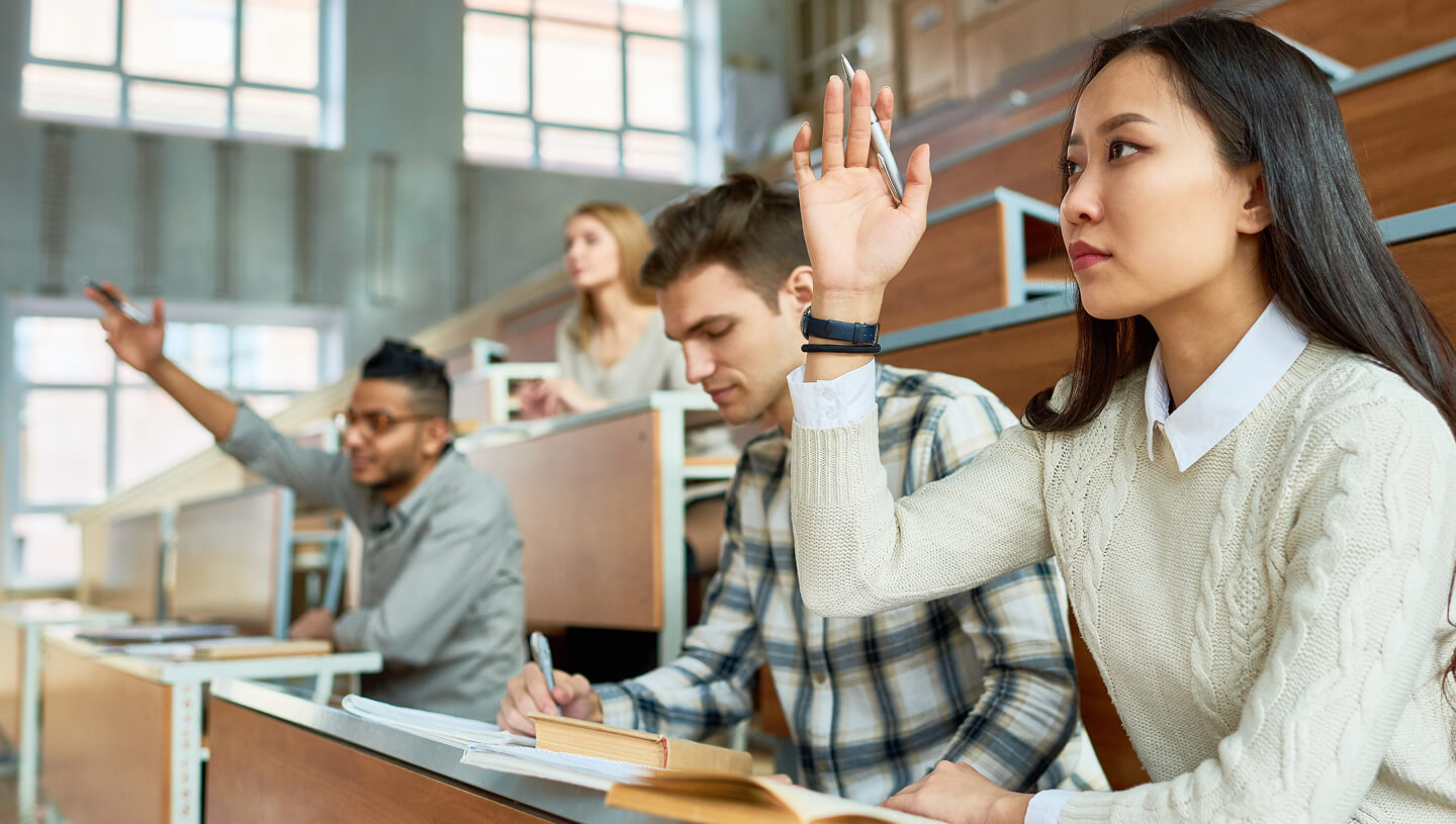 A university classroom with students seated at desks, while a student in the foreground raises her hand.