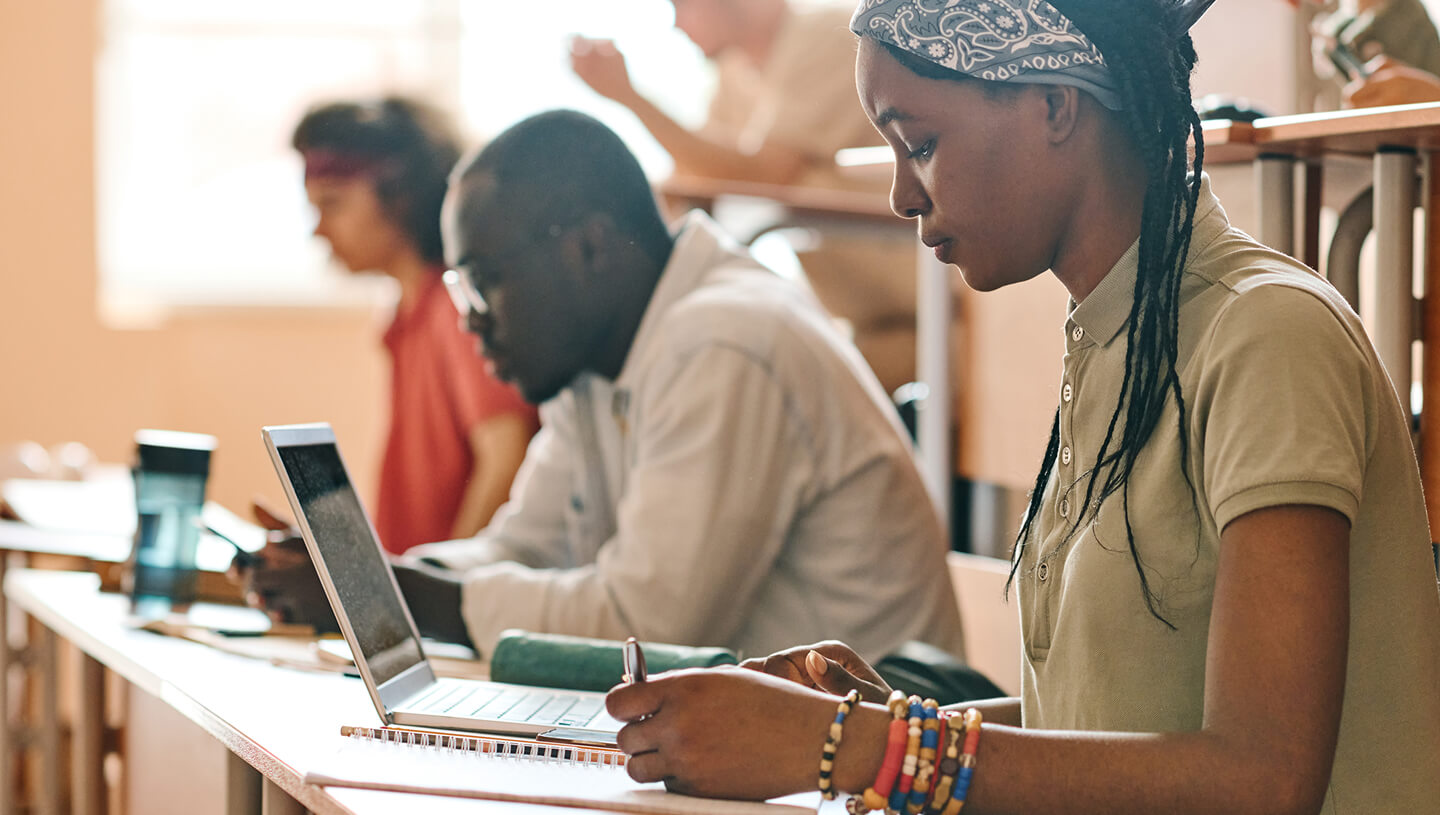 Students working in a classroom, with a student using a laptop and taking notes at a desk.